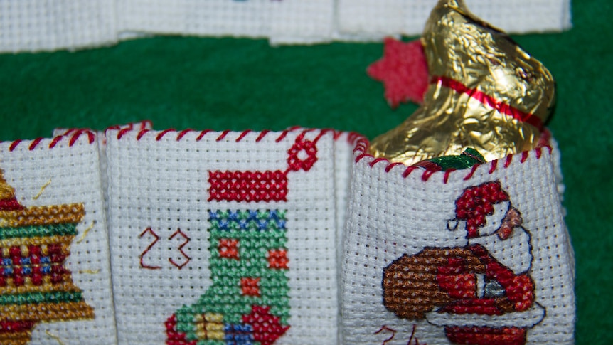 A chocolate bunny in a gold wrapper peeps out of a pocket on a cross stitched advent calendar