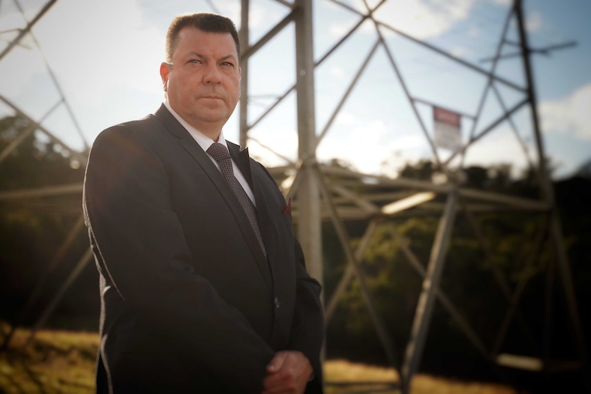 Energy analyst Marc White, wearing a dark suit and tie, stands at the base of a high-voltage power tower