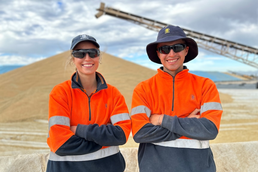 A young woman and a young man, both wearing sunglasses, with their arms crossed standing in front of grain.