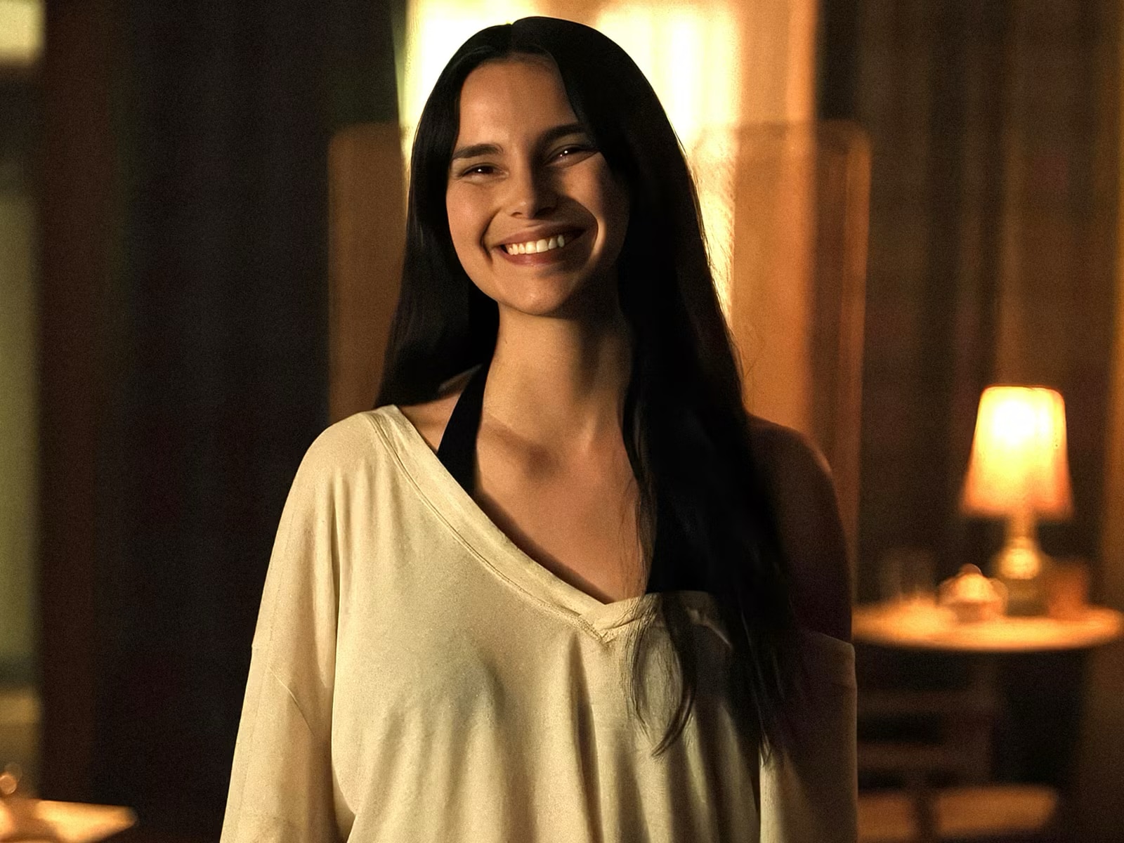 A woman with long brown hair stands in a cosy room with an unnerving smile