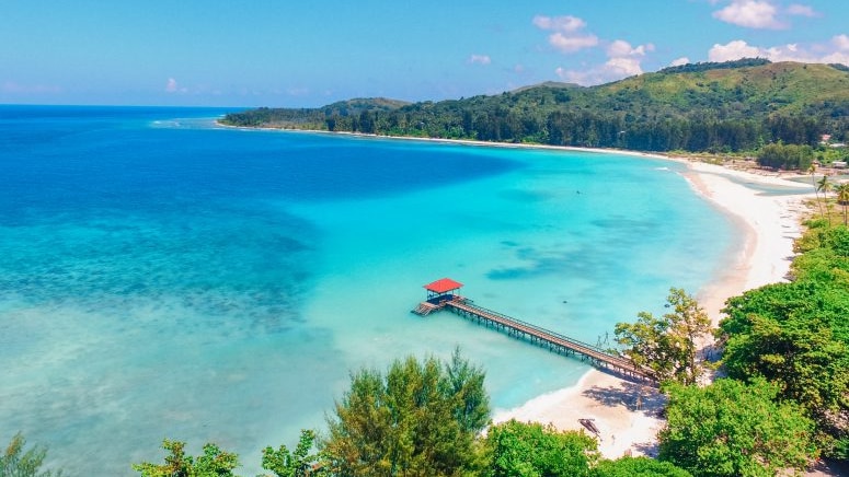 Calm turqious waters meet a thin strip of white sand that leads to a a verdant horseshoe shaped stretch of land.