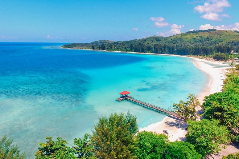 Calm turqious waters meet a thin strip of white sand that leads to a a verdant horseshoe shaped stretch of land.