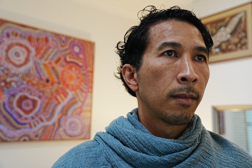 A man with black hair and a blue scarf standing in front of Indigenous art work.