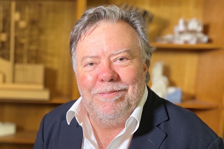 A man with grey hair and a beard wearing a blue jacket in front of a shelf with models on it