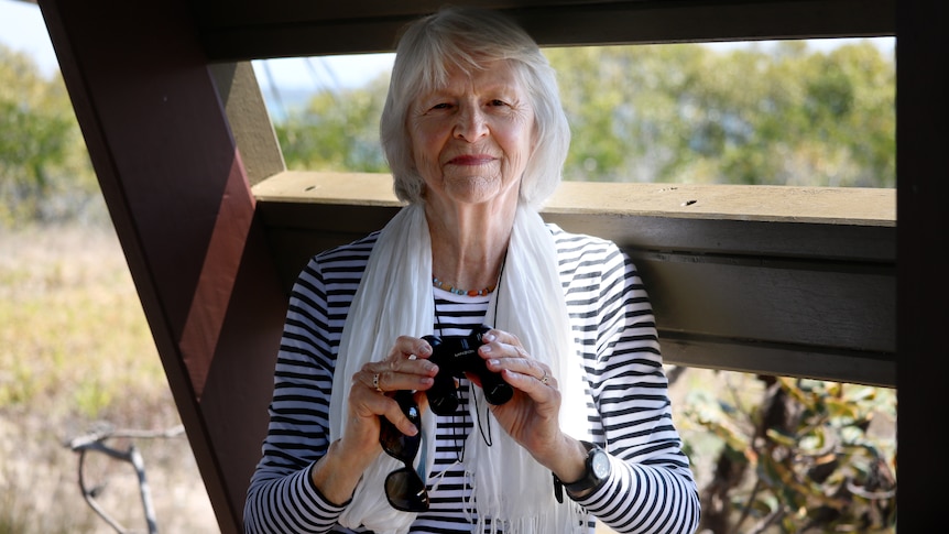 A woman with grey hair holding a pair of binoculars and sunglasses, inside a birdwatching hut.