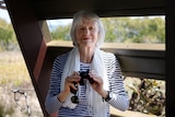 A woman with grey hair holding a pair of binoculars and sunglasses, inside a birdwatching hut.