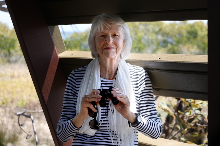 An older woman with grey hair stands inside a bird watching hut, holding binoculars and her sunglasses as she smiles.