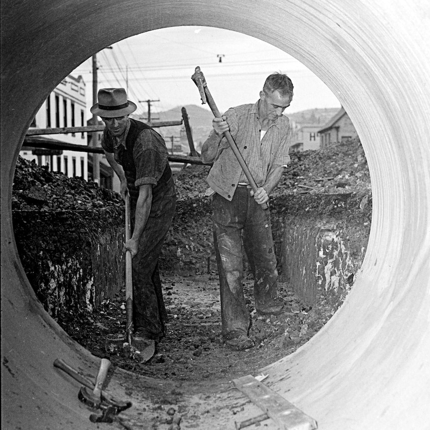 A black and white image of two men in the 1940s using tools to dig inside a large cement pipe