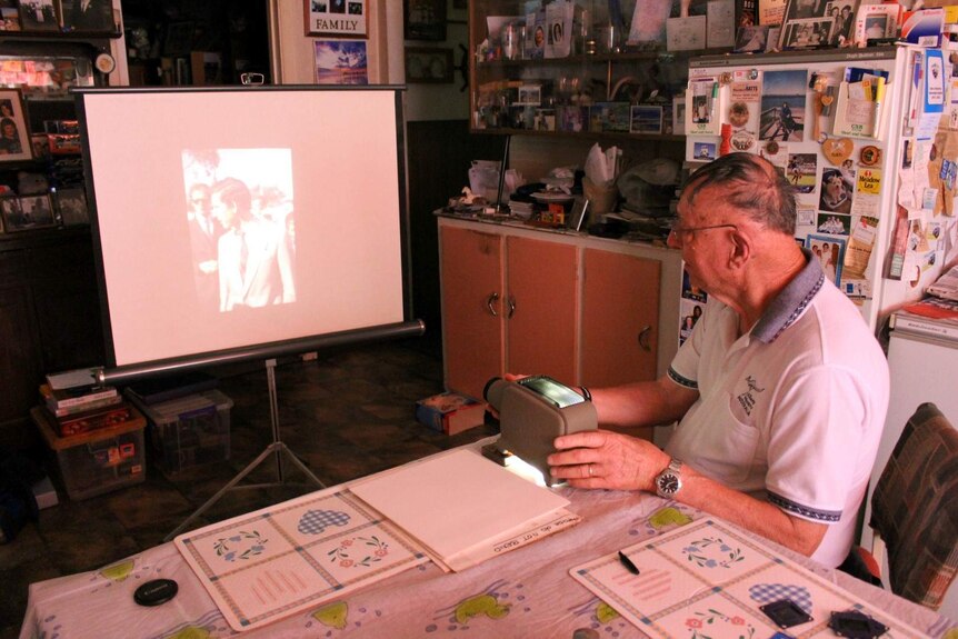 Alan looks at a photo of Prince Charles on a projector at his kitchen table. The room glows red.