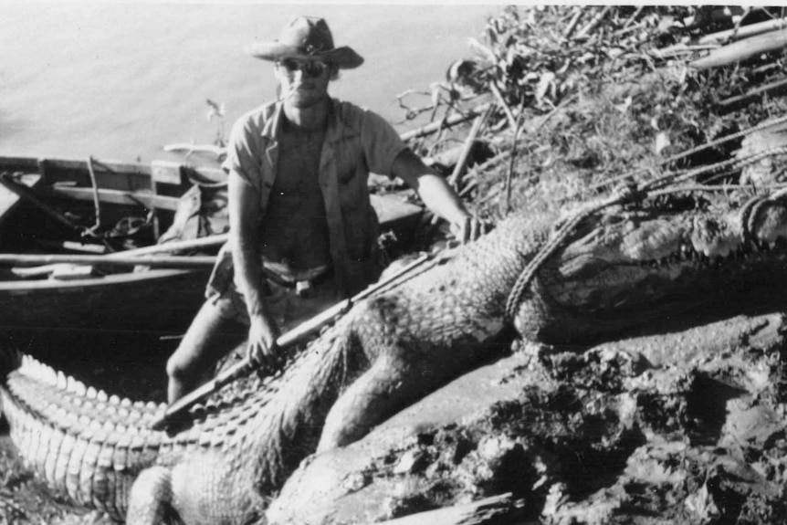 Black and white photo of man wearing hat and sunglasses sitting on a riverbank behind a tied up crocodile.