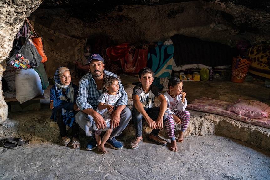 A man sits on a natural stone step inside a cave, with four children sitting next to and on his lap