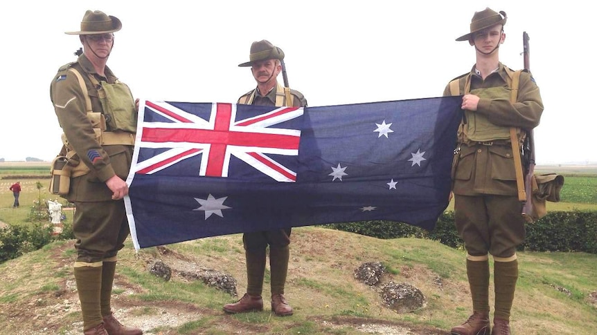 Three men face the camera wearing army uniforms and an Australian flag