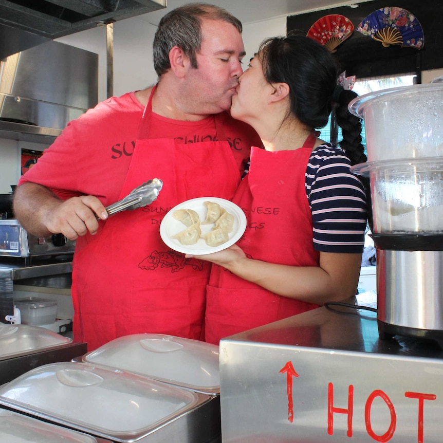 An Australian man and a Chinese woman wearing matching red aprons and holding Chinese dumplings kiss inside a dumpling cart