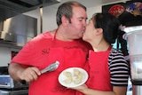 An Australian man and a Chinese woman wearing matching red aprons and holding Chinese dumplings kiss inside a dumpling cart