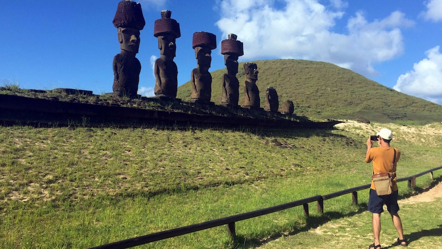 A man in an orange T-shirt and white cap takes a photo of a row of monuments on a sunny day.