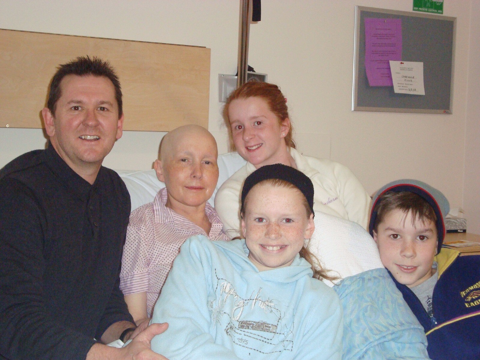 A man and three children gather around a hospital bed with a bald woman smiling