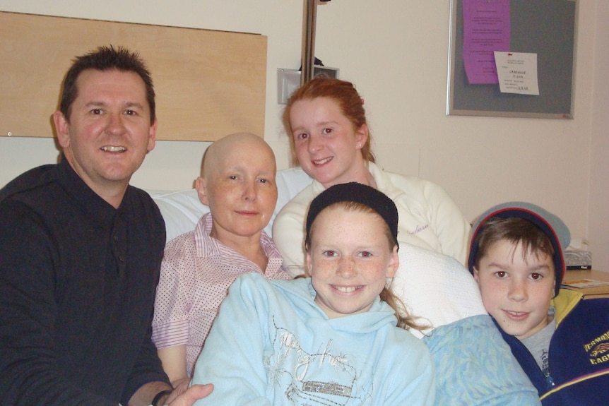 Un homme et trois enfants se rassemblent autour d'un lit d'hôpital avec une femme chauve souriant