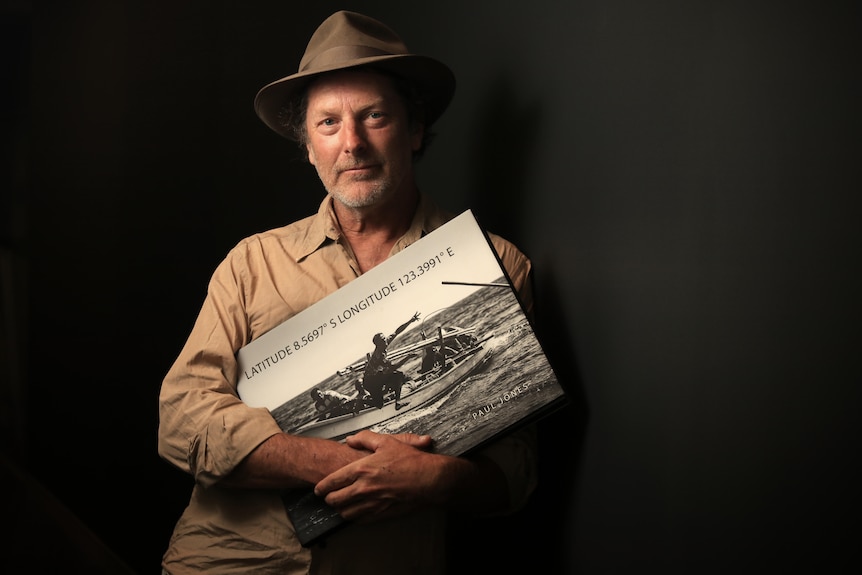 A man stands against a dark backdrop and holds a coffee-table-style book.