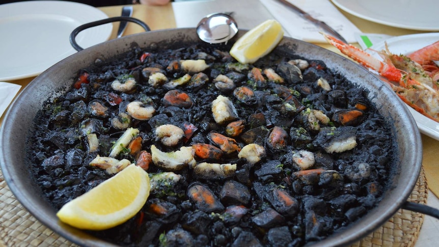 Paella negra pan with prawns and seafood, two lemon wedges rest on the edge of the pan, which sits on a restaurant table.
