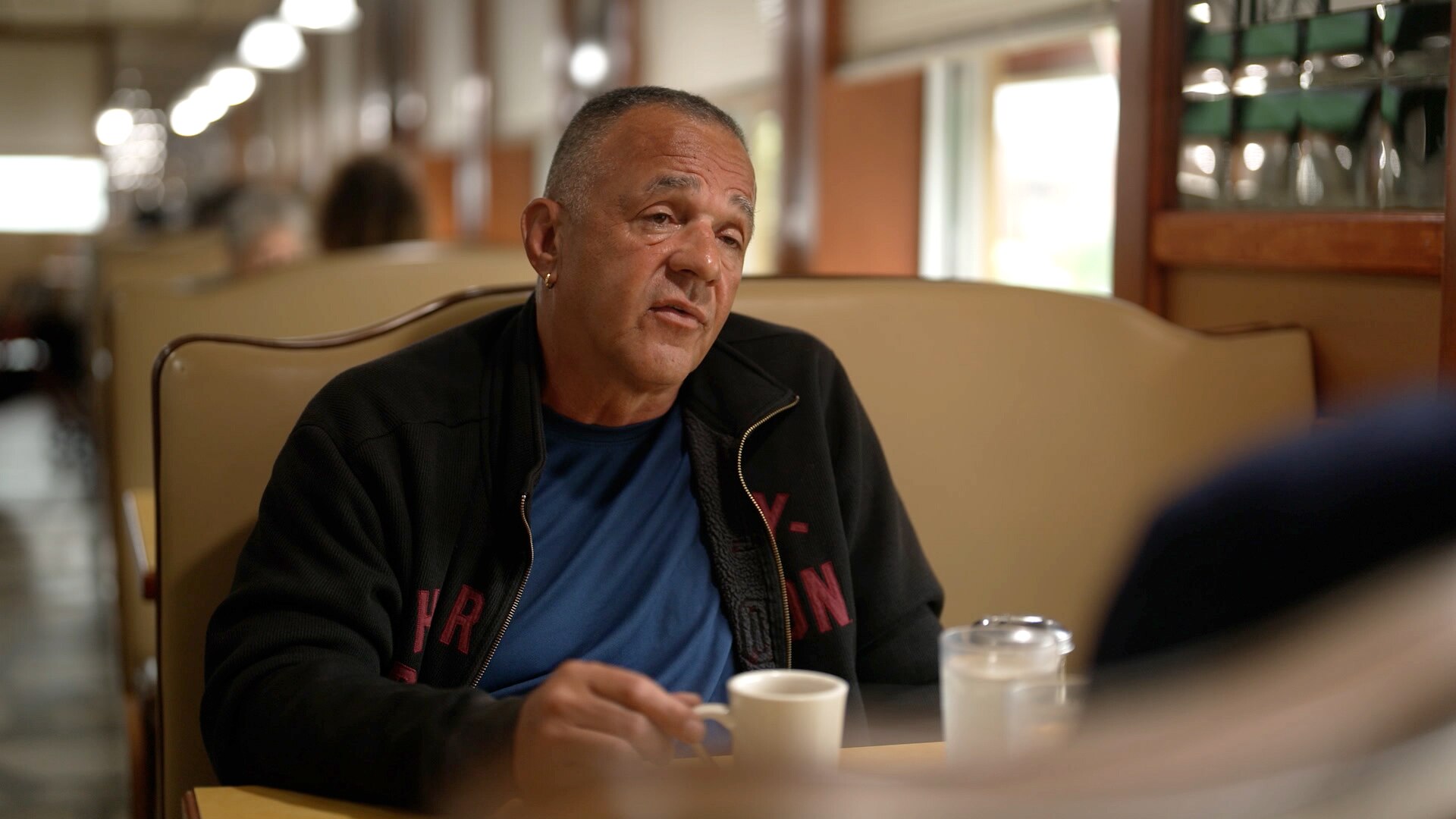A man sits in a booth at a diner, holding a coffee cup. He is looking ahead with a contemplative expression.