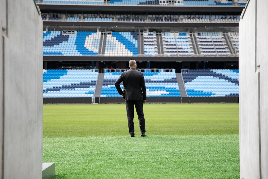 A man stands and looks over an empty football stadium, looking up at the stands with back turned 