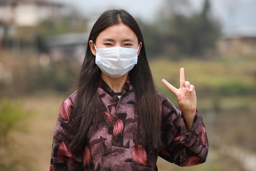 A young Bhutanese woman with straight black hair on a face mask makes a peace sign 