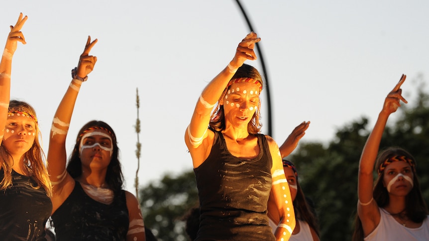 Top of body shot of five female Noongar dancers on stage at dusk, right arms raised and index and middle fingers pointed.