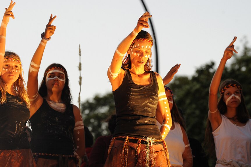 Top of body shot of five female Noongar dancers on stage at dusk, right arms raised and index and middle fingers pointed.