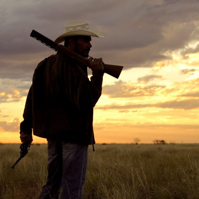 Actor Aaron Pederson wearing a wide-brimmed hat and with a shotgun over his right shoulder, in an Australian bush landscape.