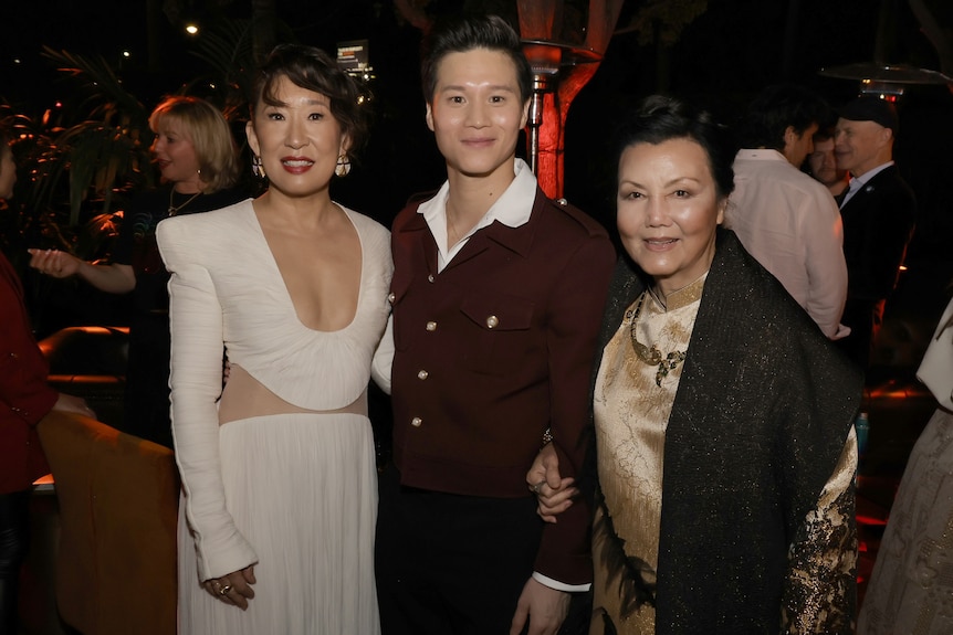Three Asian actors pose at an event: a middle-aged woman in a white dress, a young man in an open-collar suit and an older woman
