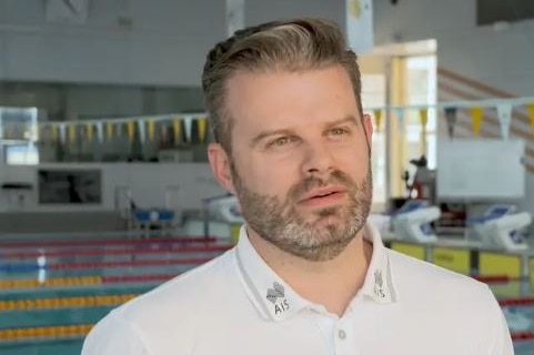 A man in white polo shirt with facial hair and neutral expression stands with a pool in the background.