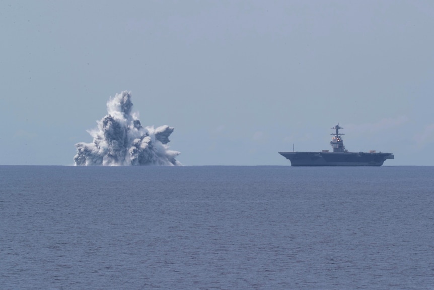 A sea level view of a huge cascade of water exploding into the air nearby an aircraft carrier.