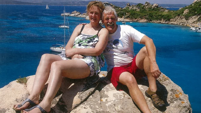 Jacinta Bohan (left) and Trevor Salvado sit on a rock with the ocean behind them, smiling at the camera.