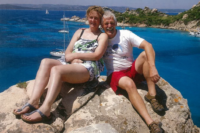 Jacinta Bohan (left) and Trevor Salvado sit on a rock with the ocean behind them, smiling at the camera.