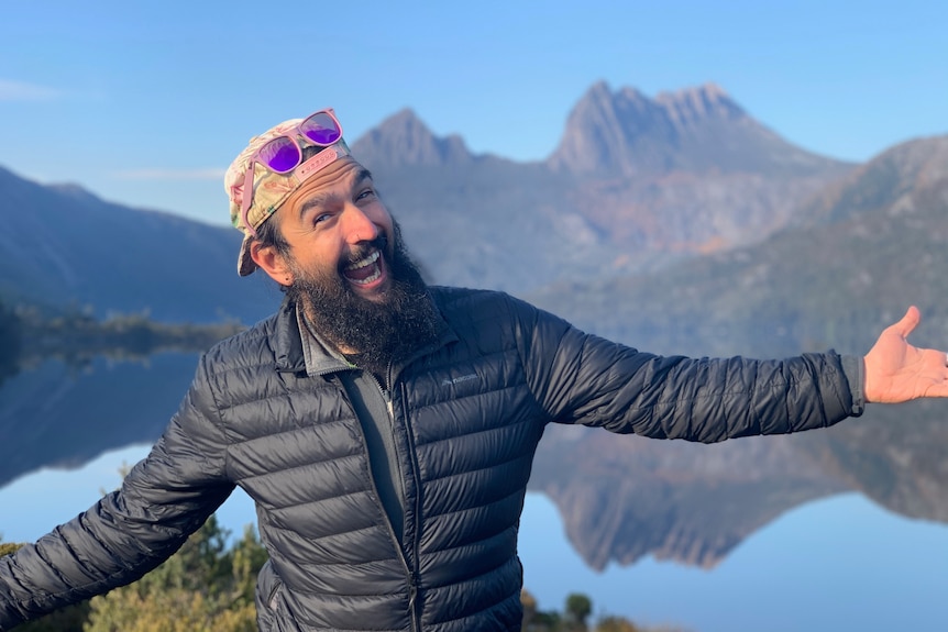Man wearing hiking attire, pink sunglasses and hat with arms outstretched at a viewing point with Cradle mountain in background.