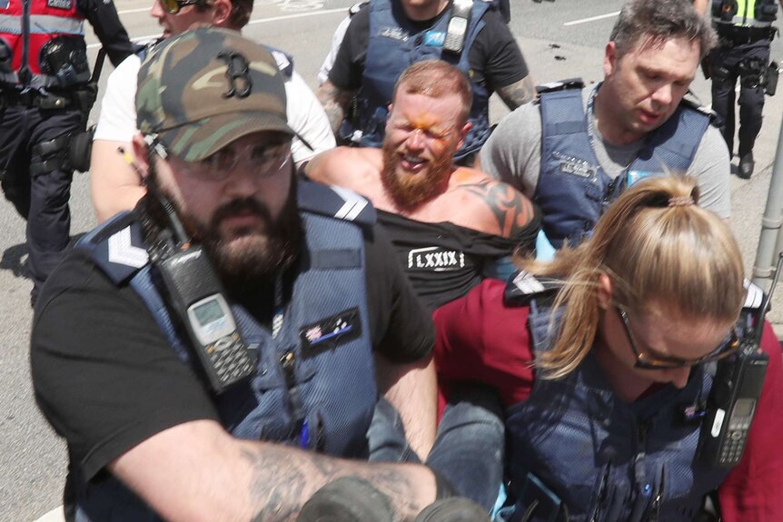 Four police officers wearing vests and plain clothes escort a man who has orange pepper spray on his face.