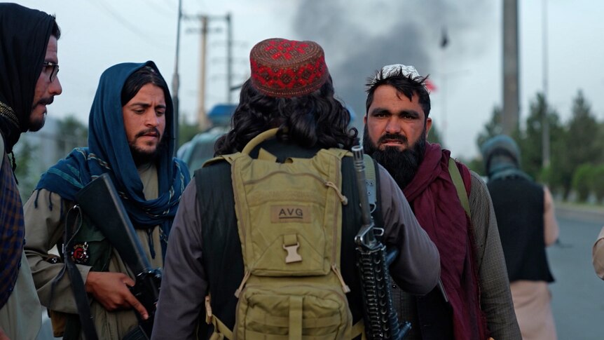 A group of Taliban fighters stand in a street. One is holding a machine gun, one is looking sternly at the camera.