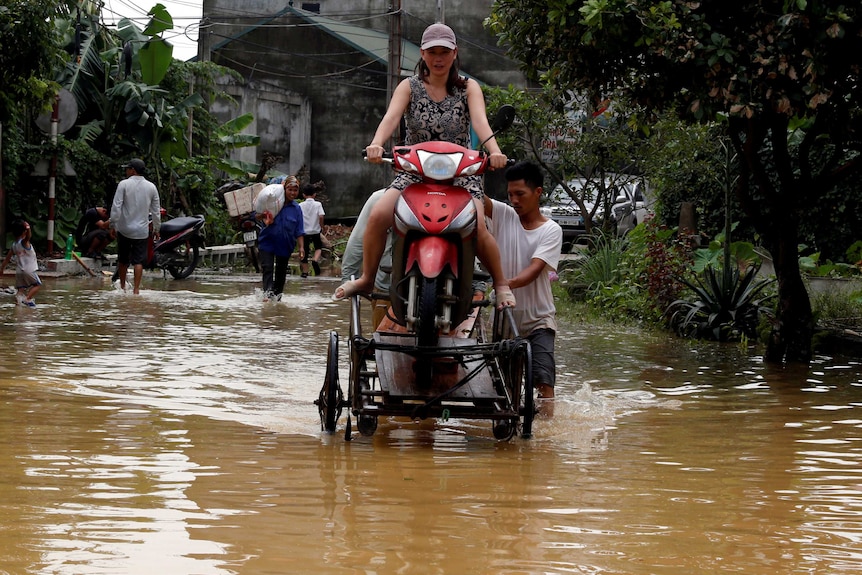 A woman sits on a scooter as men push her through floodwaters.