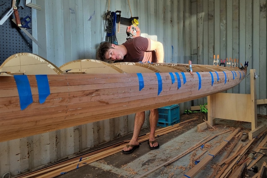 A man bending over while looking along the length of a kayak he is making from timber.