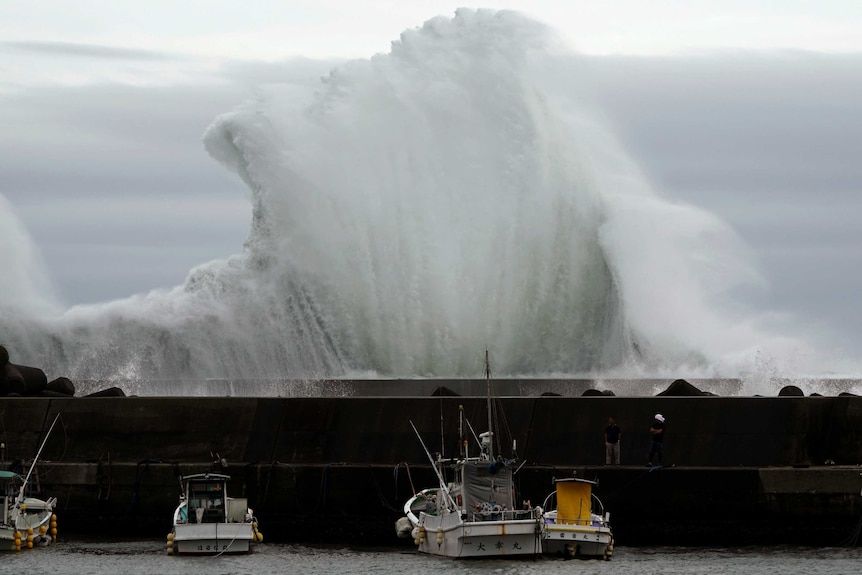 A massive wave crashes into a wall shielding boats at a port.