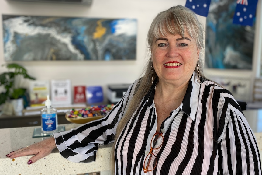 A smiling woman in a black and white striped blouse stands in front of a hotel welcome desk