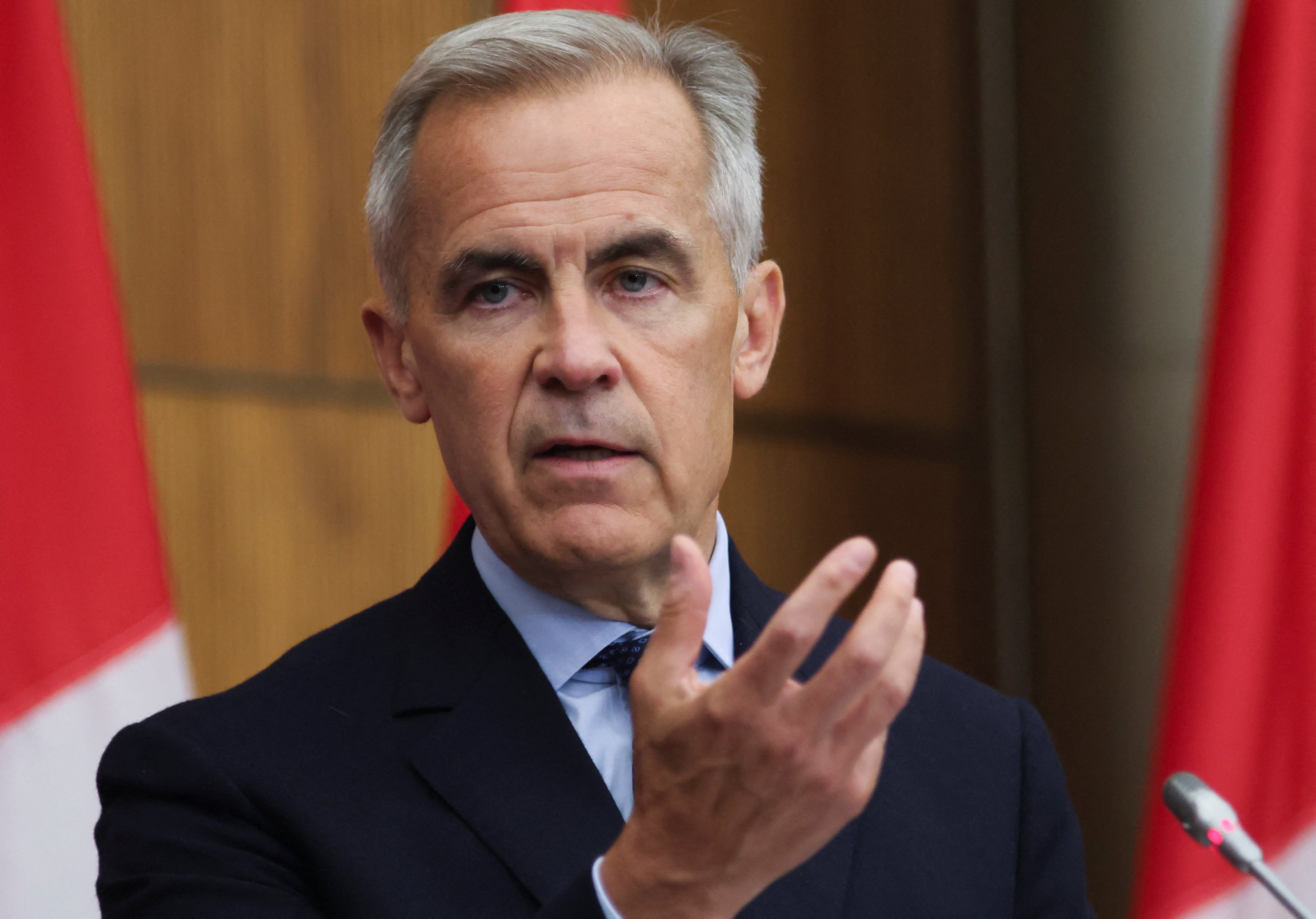 A man in a black suit gestures in front of two Canadian flags