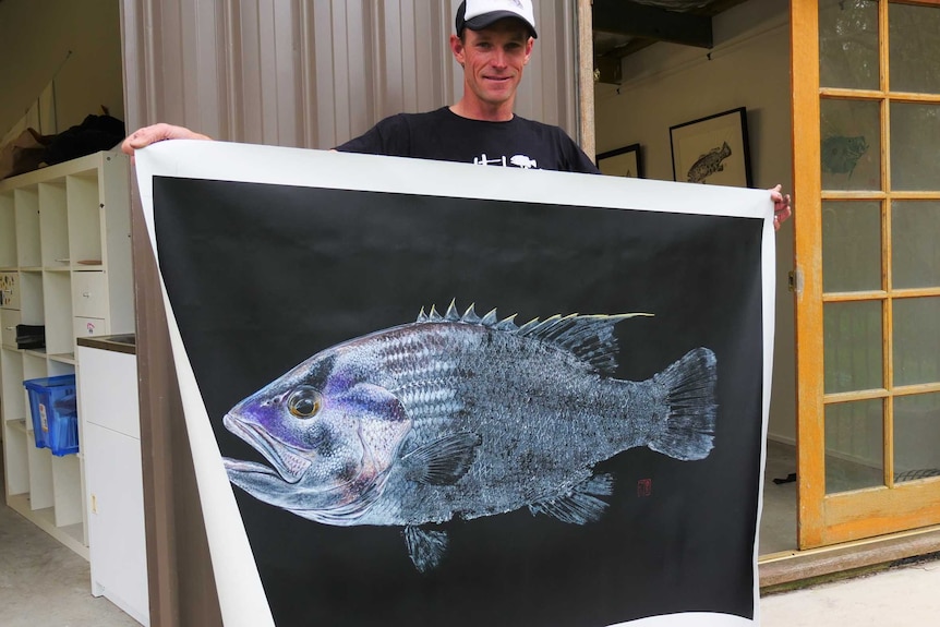 A man stands outside his studio holding a large canvas with a fish.