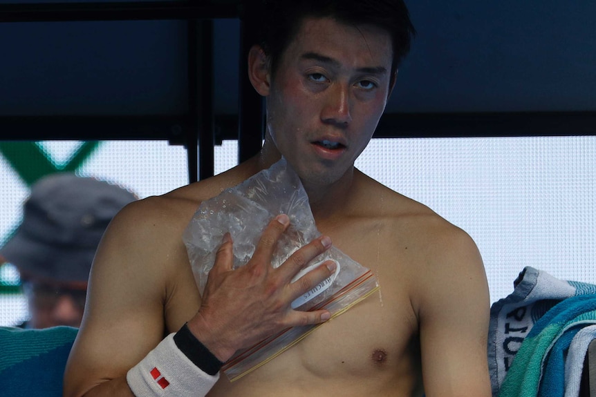 A man sits in the shade with his shirt off, pressing a clear plastic bag filled with ice on his chest