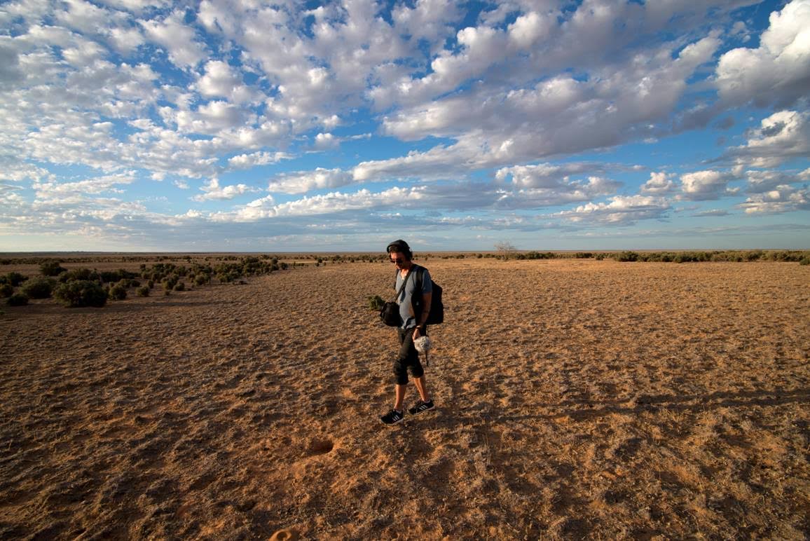 An Aboriginal man with audio recording equipment stands in a dessert, fluffy clouds in the sky