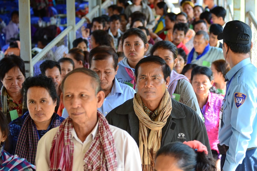 A large group of people, many wearing checked scarves, walk past a security guard.