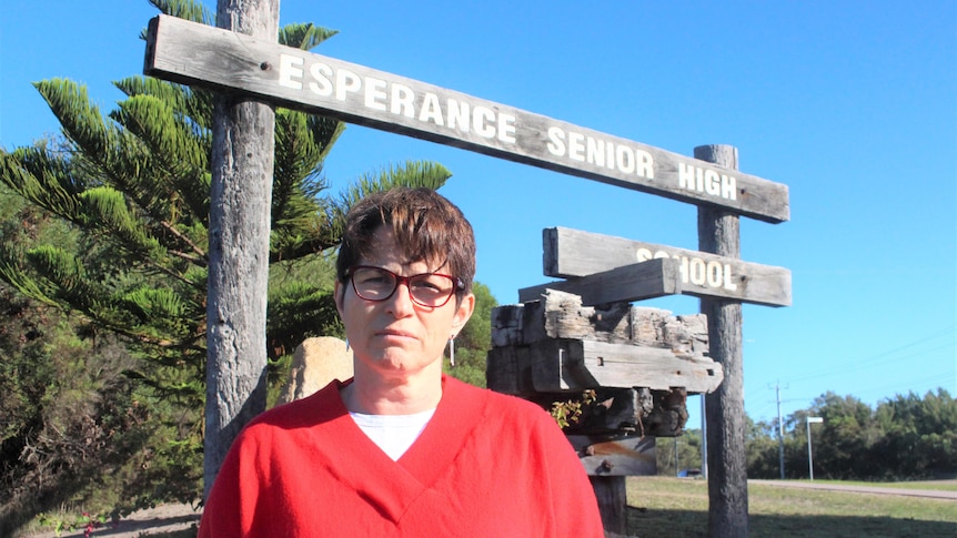 She has short brown hair and wears a bright red jumper and stands, frowning, in front of a wooden sign with the school's name on