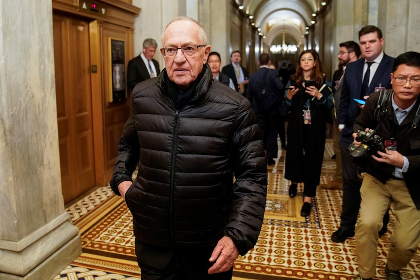 Alan Dershowitz in a black puffer jacket walking through the halls of Congress amid photographers