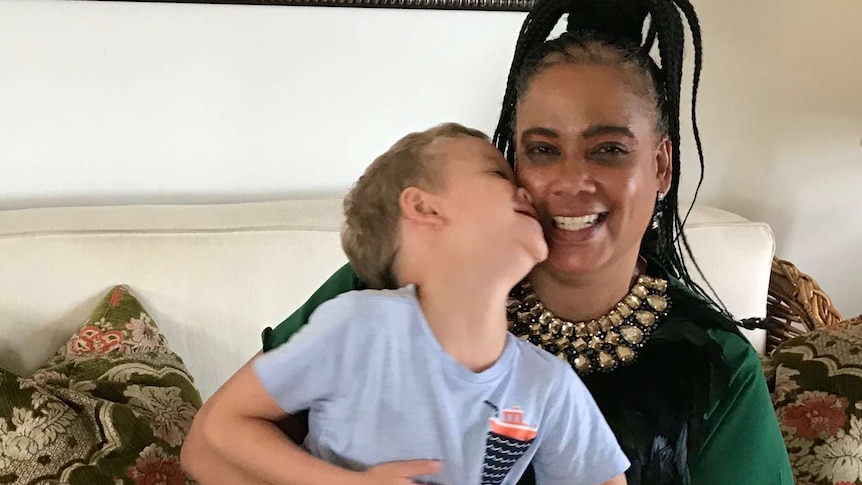 A woman sit on a lounge smiling while her son sits on her lap pressing his face against the side of hers