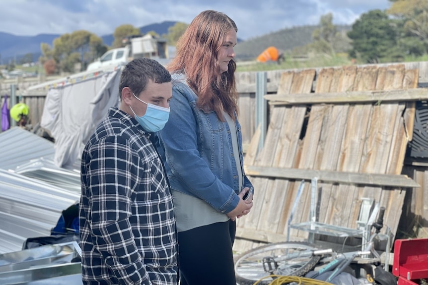 A man and a woman stand in a messy backyard near a fence.